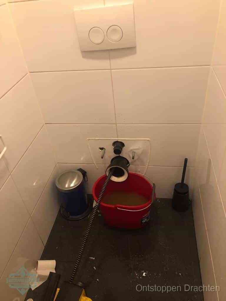 Een ontstopte wand-wc met verwijderde toiletpot waar een ontstoppingsveer in de wanduitgang verdwijnt, het achterhalen waar de verstopping zit in Drachten