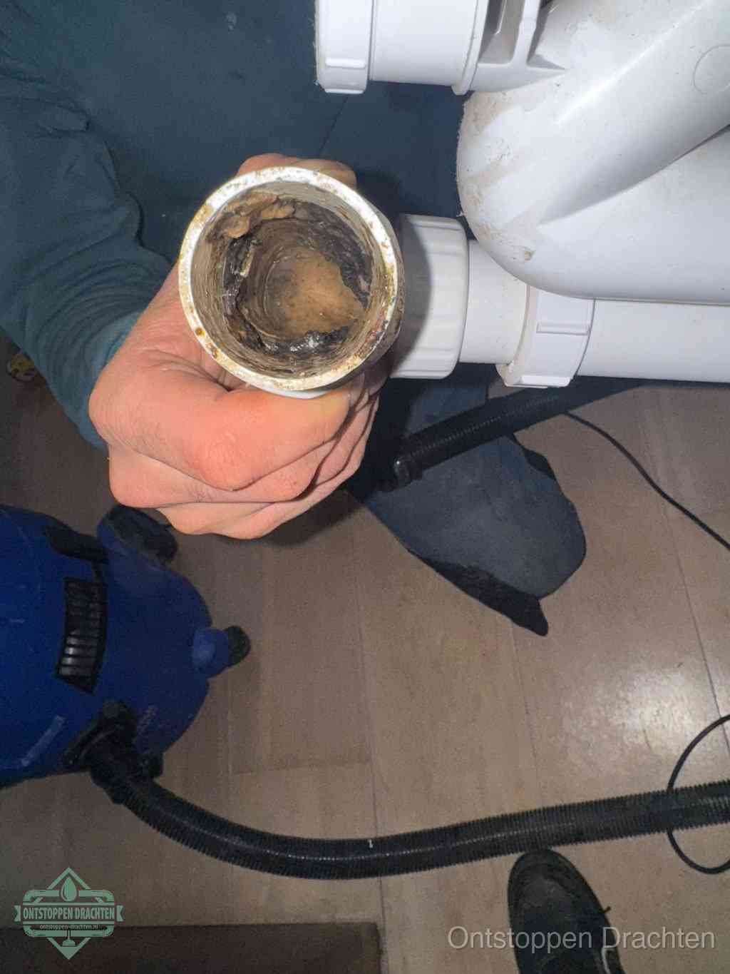 Een hand houdt een losgekoppeld PVC-afvoerbuisstuk vast dat van binnen volledig is dichtgeslibd met vet, vuil en aanslag, de meest voorkomende oorzaak van een verstopte afvoer in Drachten