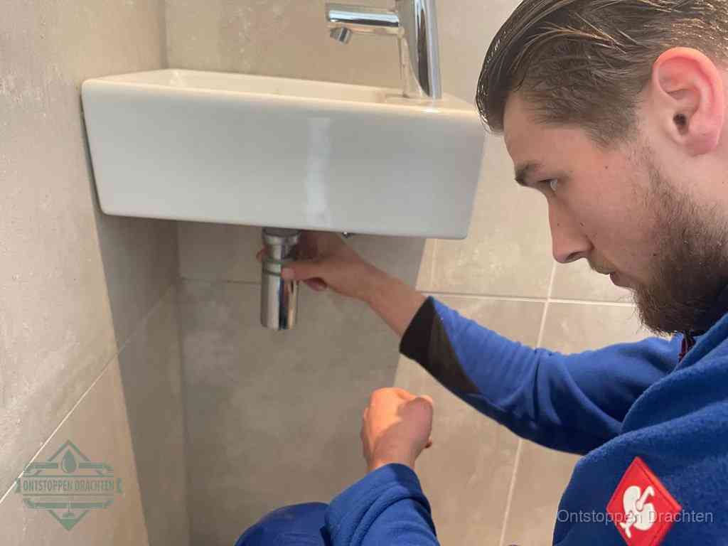 Een monteur draait met de hand de zwanenhalssifon onder een fonteinwasbak in de toiletruimte los om de bron van rioollucht in Drachten te vinden