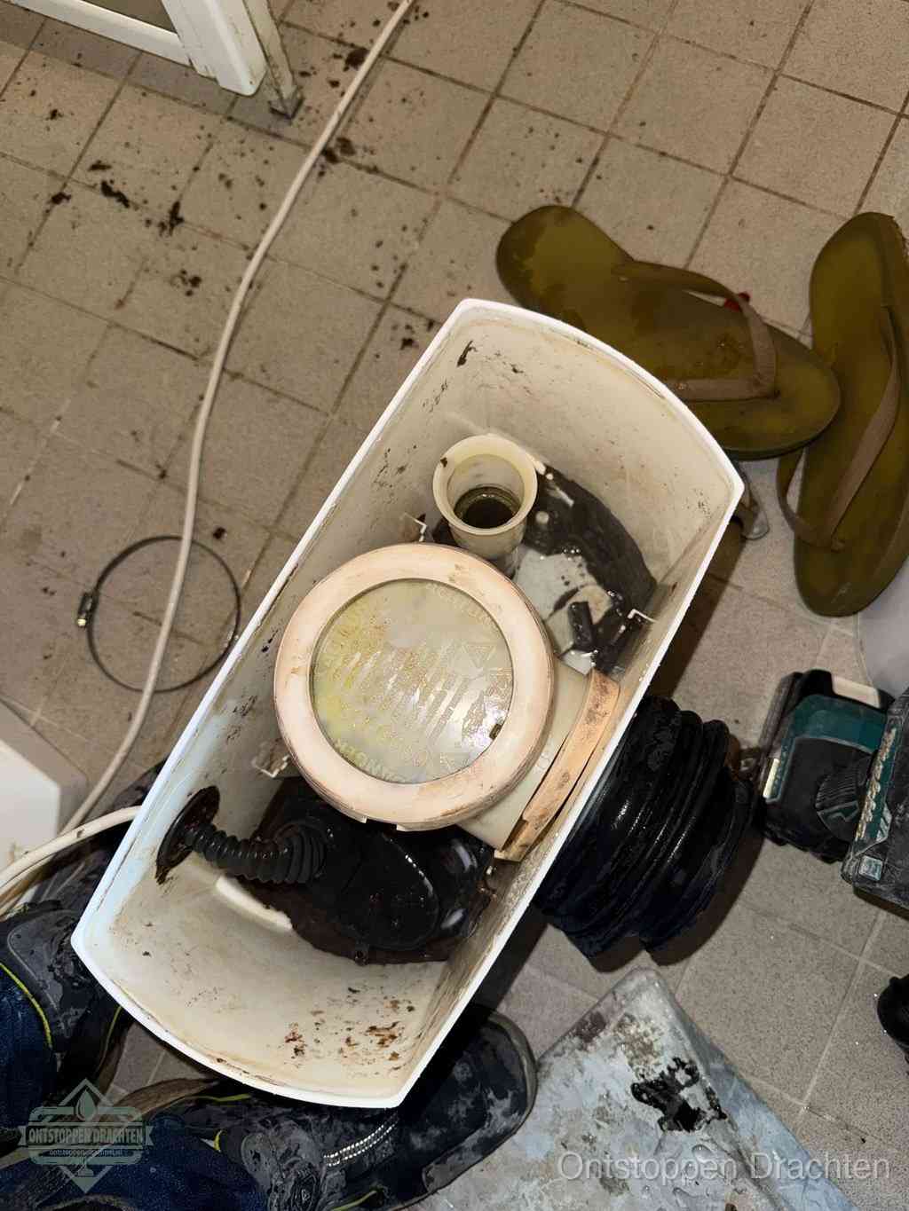 Een gedemonteerde toiletpot op de vloer met ontstoppingsveer en gereedschap ernaast, het verwijderen van een vastzittend voorwerp uit de wc in Drachten