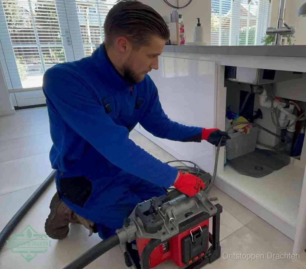 Een monteur met rode handschoenen knielt in een keuken en voert een elektrische ontstoppingsveer in tijdens het professioneel afvoer ontstoppen in Drachten